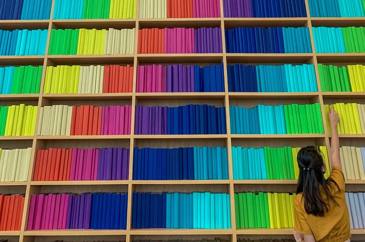 A woman in an orange shirt reaching up to grab a yellow book from a large bookshelf with all the books arranged perfectly by color.