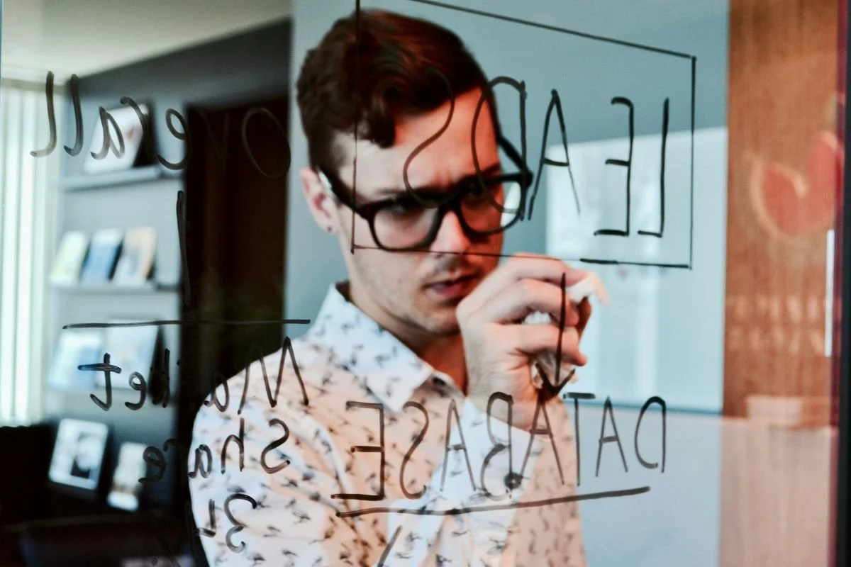 A young man with glasses writing on a glass board with a marker, with words like 'LEADERSHIP' and 'DATASAFE' visible.