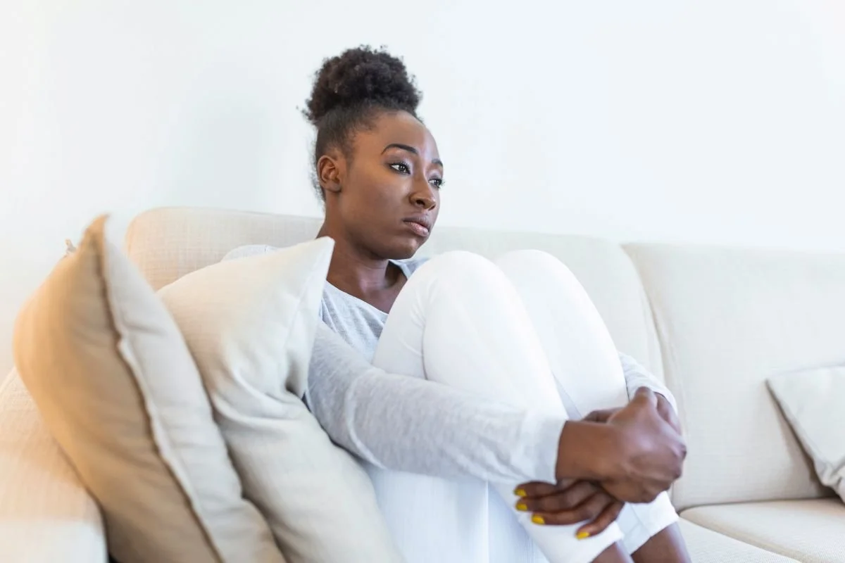A woman sitting on a light-colored sofa, hugging her knees, with a pensive or worried expression on her face, in a room with white walls.