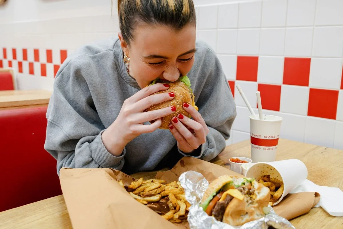 A woman with blonde hair in a bun, wearing a gray sweatshirt, rapidly eating a large burger at a fast-food restaurant. The table has a tray with fries, a cup with straws, a small container of sauce, and another wrapped sandwich.