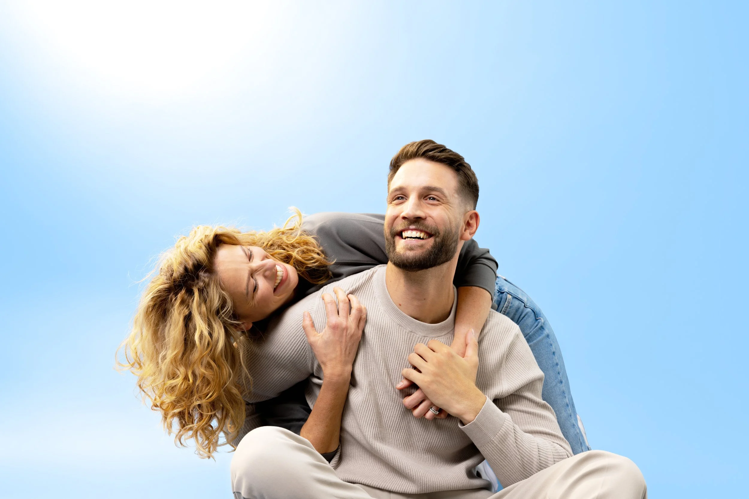 A smiling couple enjoying outdoor leisure time against a clear blue sky, with the woman on his back and both appearing joyful.
