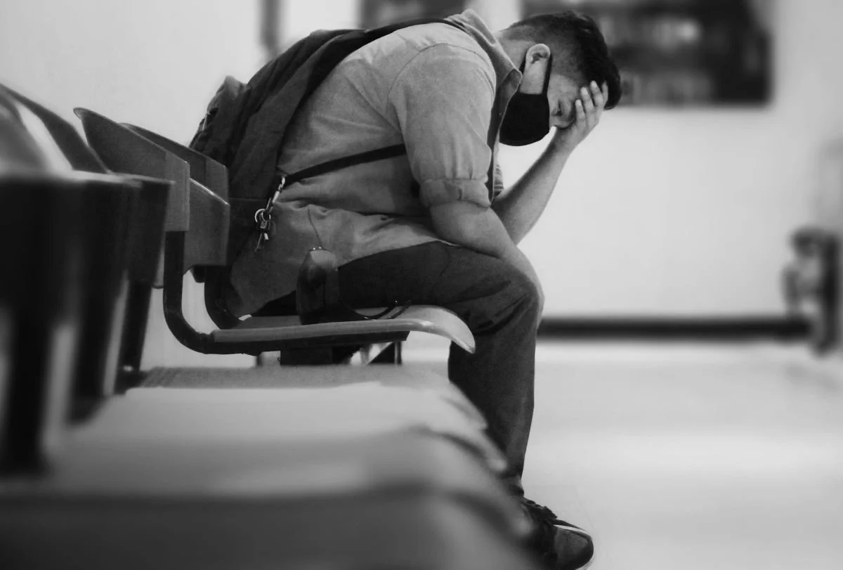 A young man sitting alone on a row of chairs in a hospital waiting area, resting with his head in his hand, appearing distressed.