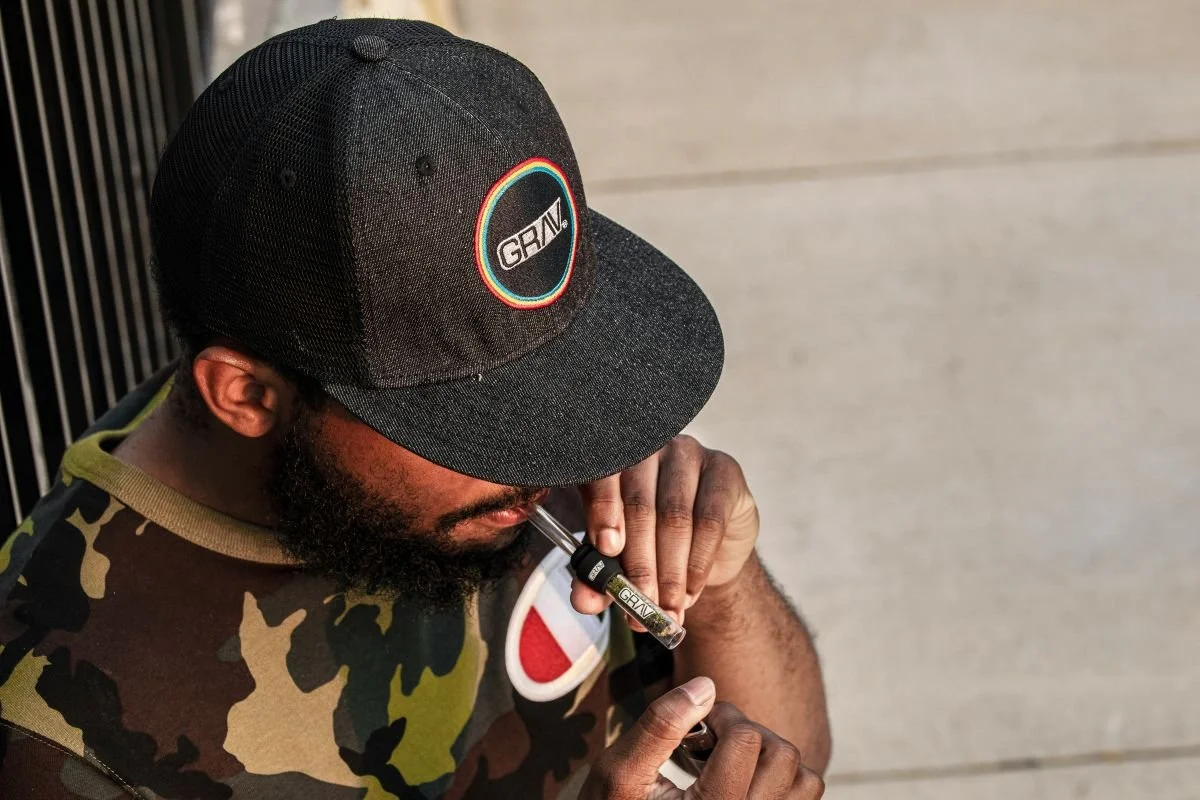 Man in camouflage shirt and black cap, holding a vape pen to his lips.