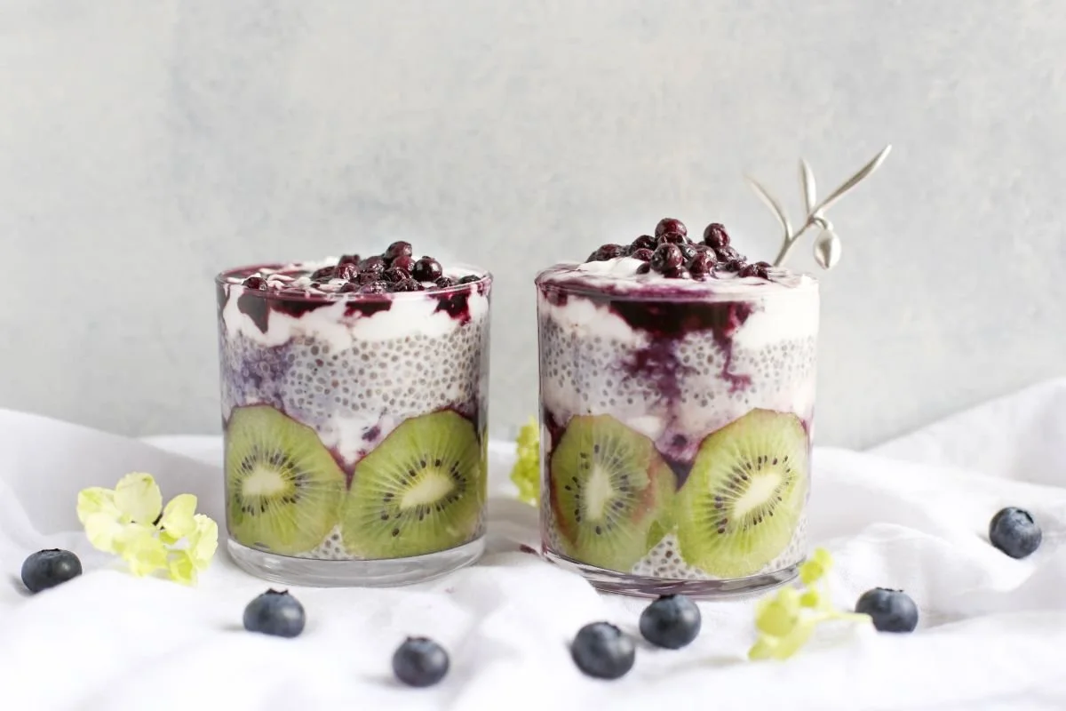 Two glasses of layered dessert with slices of kiwi, topped with berries and chia seeds, set on a white surface with scattered blueberries and small yellow flowers.