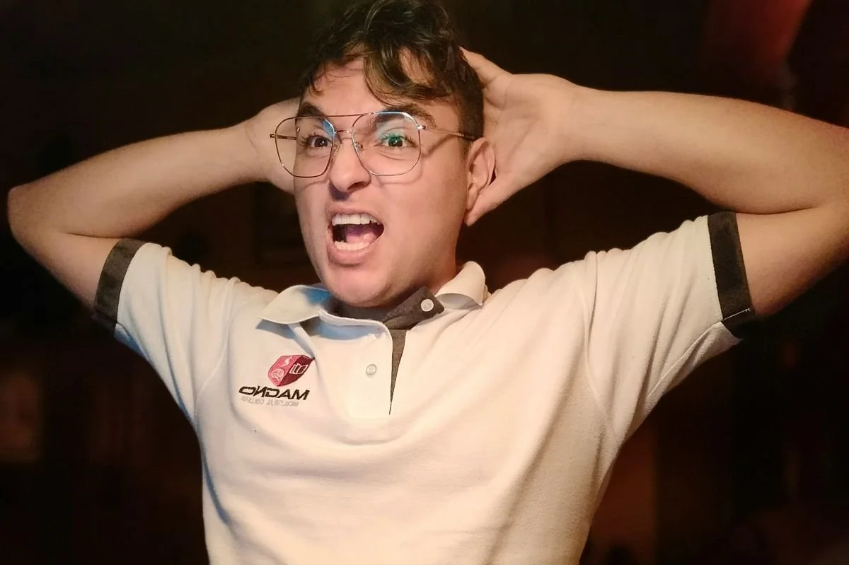 A young man with glasses and short hair, wearing a white polo shirt with black trim, appears to be yelling or surprised, with his hands behind his head in a setting with dark background.