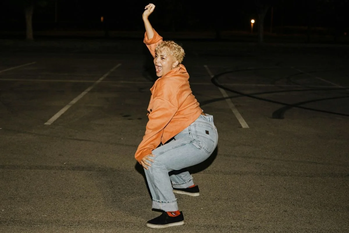 A person with curly blonde hair wearing an orange jacket, light blue jeans, and black sneakers with orange socks, laughing and twerking in a parking lot at night.