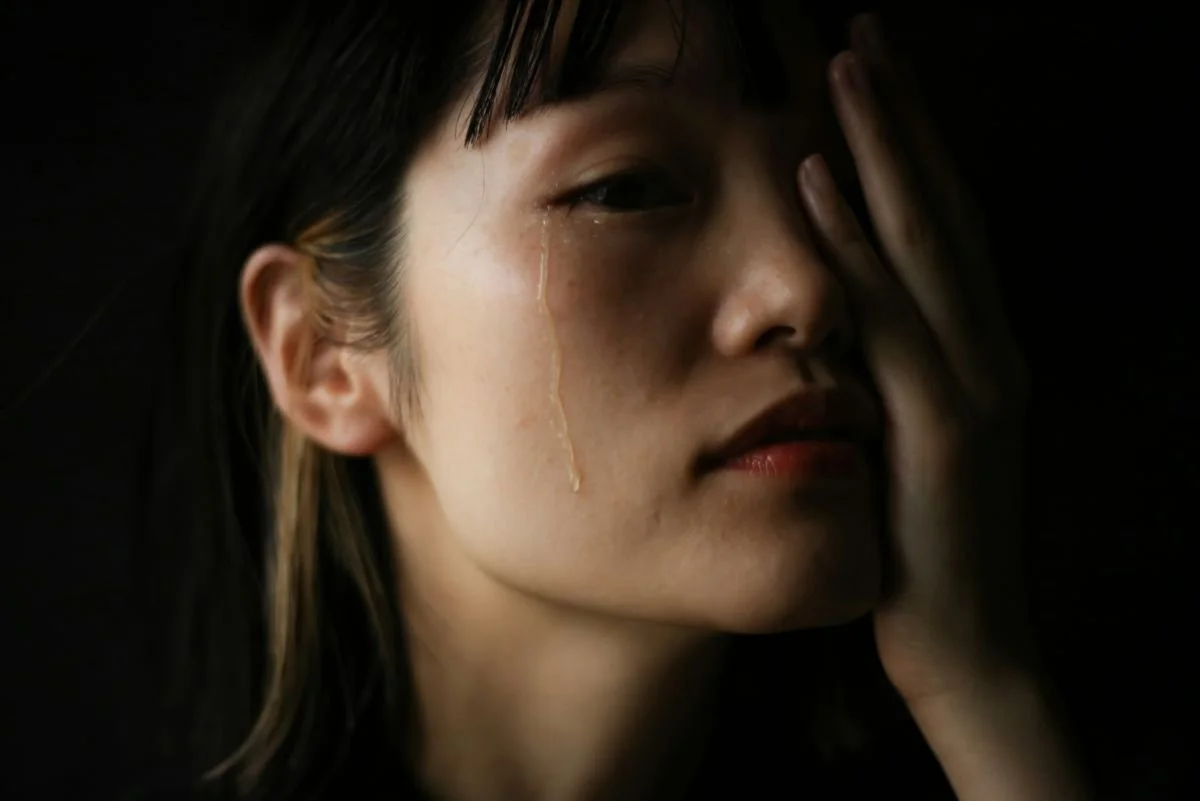 Close-up of a woman with tears running down her face, holding her face with her hand against a dark background.