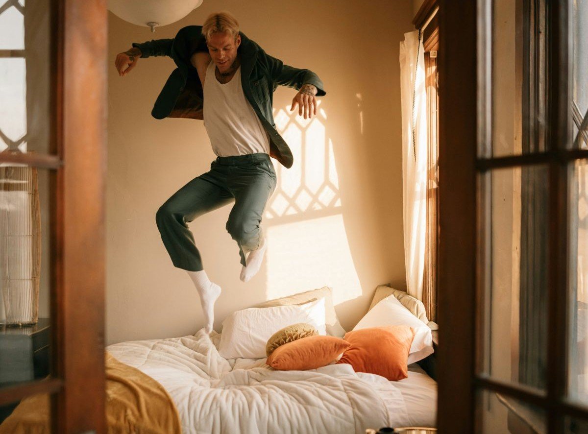 A man jumping on a bed in a sunlit bedroom, with pillows and blankets, as viewed through a window.