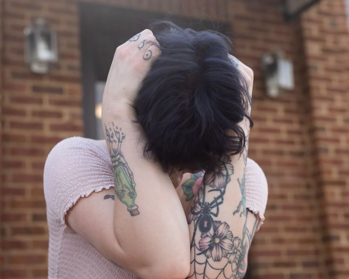 Person with short dark hair and tattoos on arms, covering face with hands, standing in front of a brick wall.