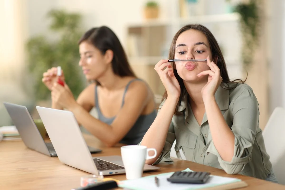 Two women sitting at a desk with laptops, one holding a lipstick and the other placing a pencil horizontally across her upper lip, mimicking a mustache.