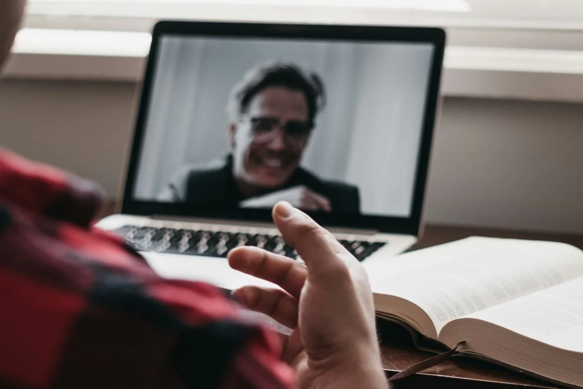 Person on a video call with a man on a laptop, open book on desk, using gesture to communicate.
