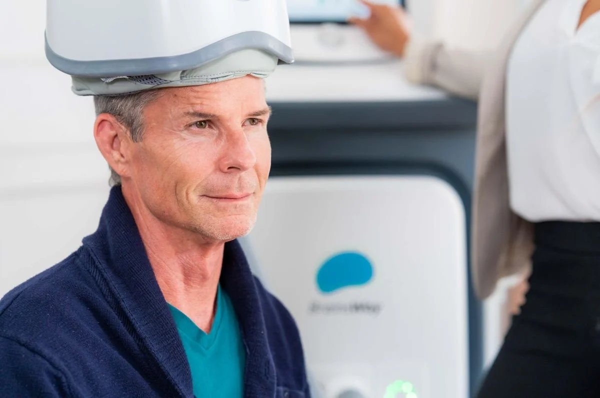 A middle-aged man receiving TMS with the helmet positioned over his head and the TMS machine visible in the background.
