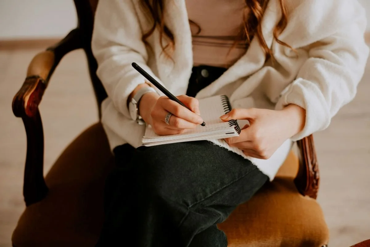 Person sitting in a wooden chair writing in a spiral notebook with a black pen.