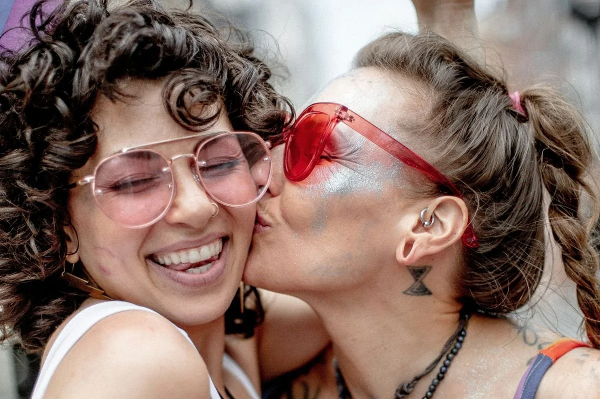 Two women smiling and hugging; one is kissing the other on the cheek in a joyful moment.