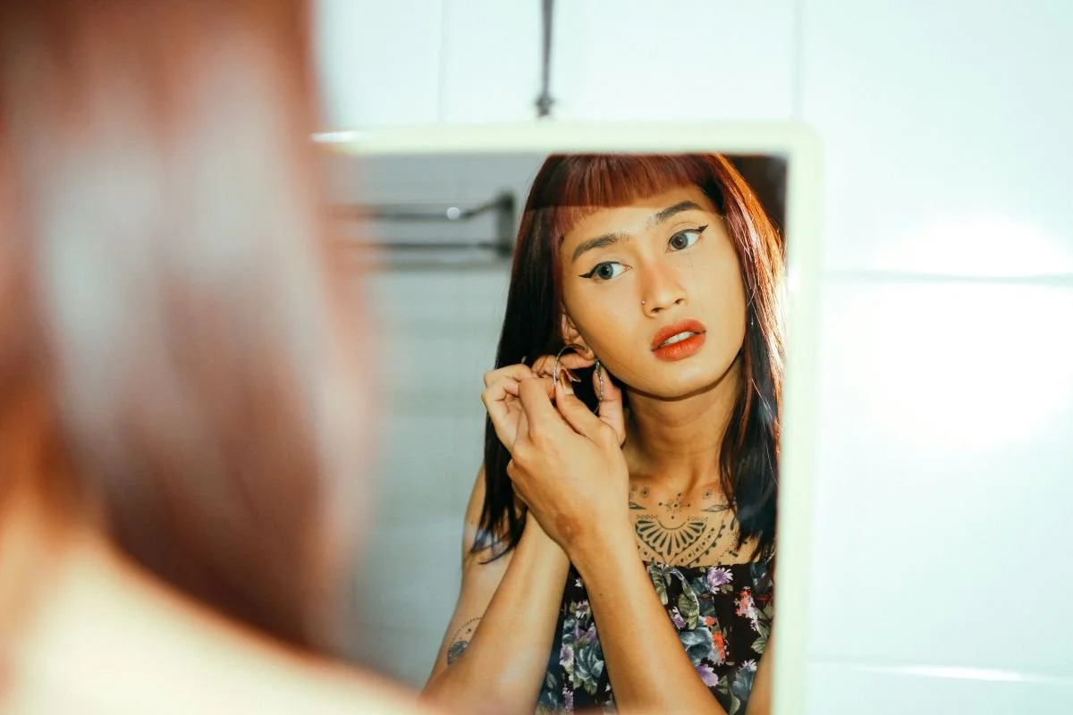 A transgender woman with black hair and blunt bangs looking in a mirror, adjusting an earring, wearing a floral top, with tattoos on her chest and neck, in a brightly lit room.