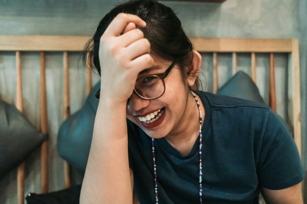 A woman with glasses smiling, holding her hand on her head, sitting in a cozy room with a wooden headboard and gray pillows in the background.
