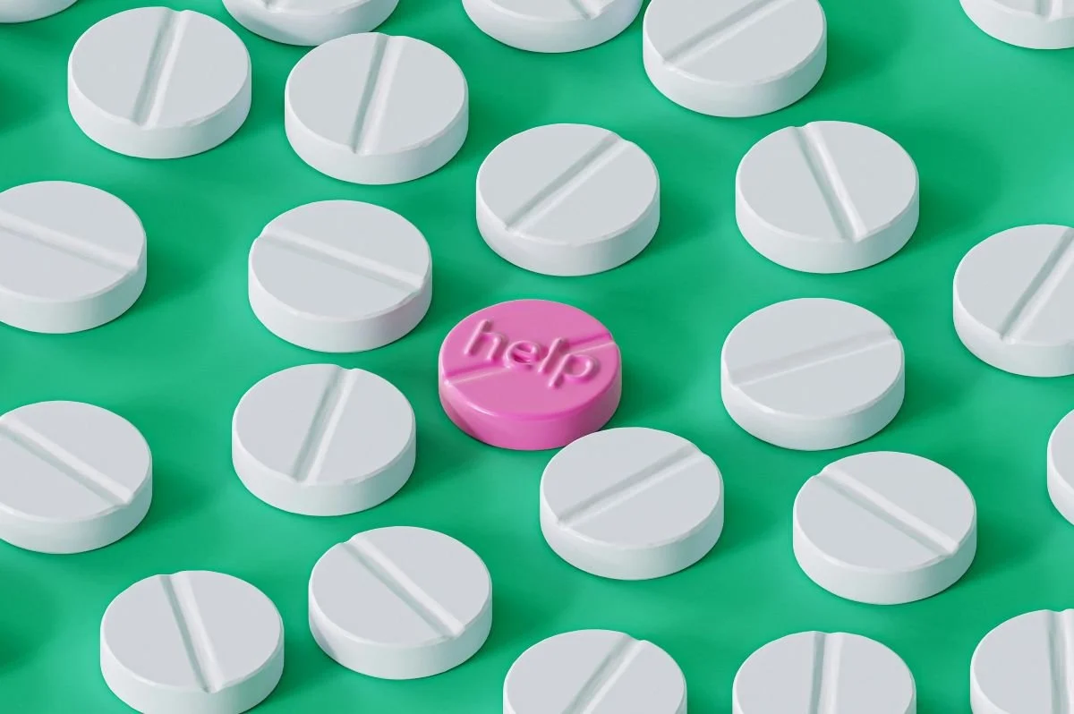 A pink pill with the word 'help' on it surrounded by many white round pills on a green background.