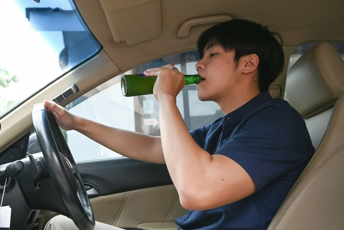 A young man sitting in the driver's seat of a car, drinking from a green beer bottle.