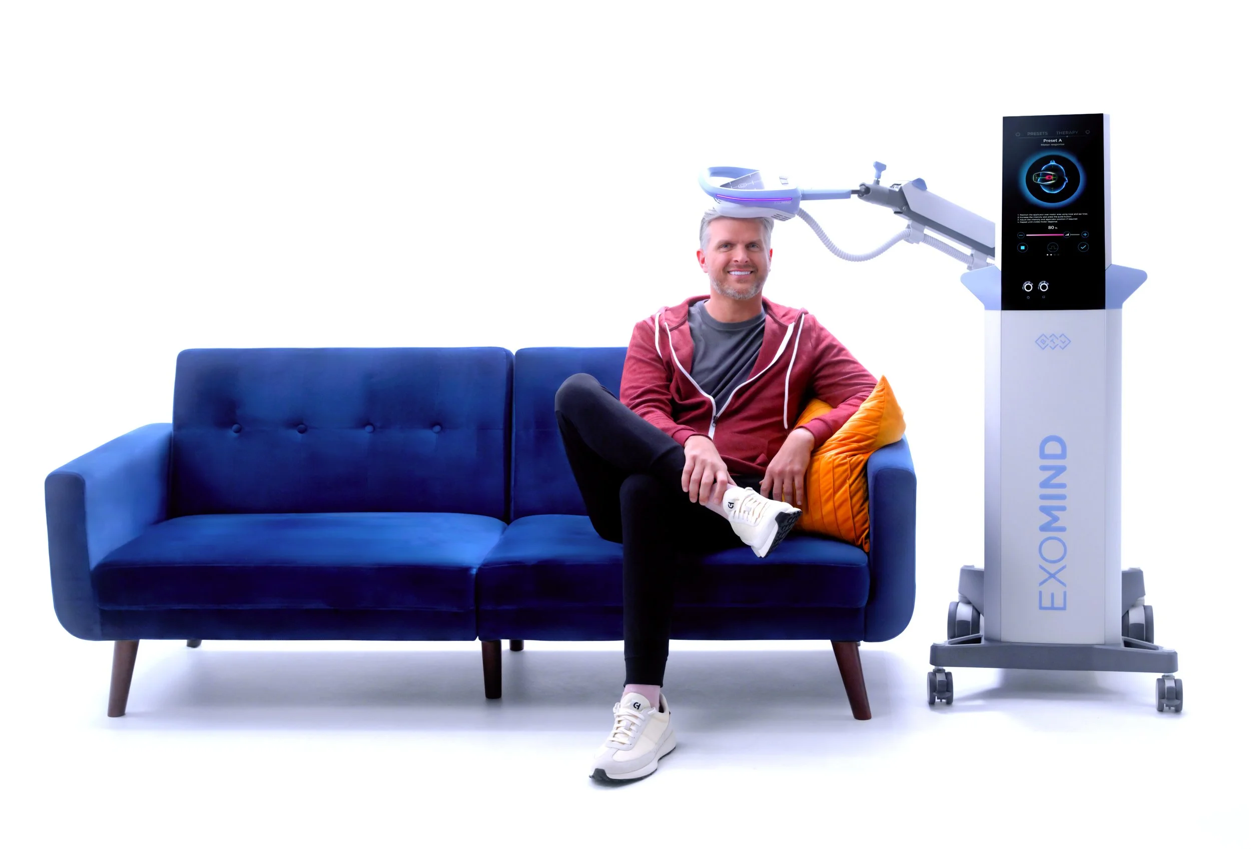 A man sitting on a blue couch with a yellow pillow, smiling, next to a futuristic medical device labeled 'EXOMIND' with a robotic arm extending over his head.