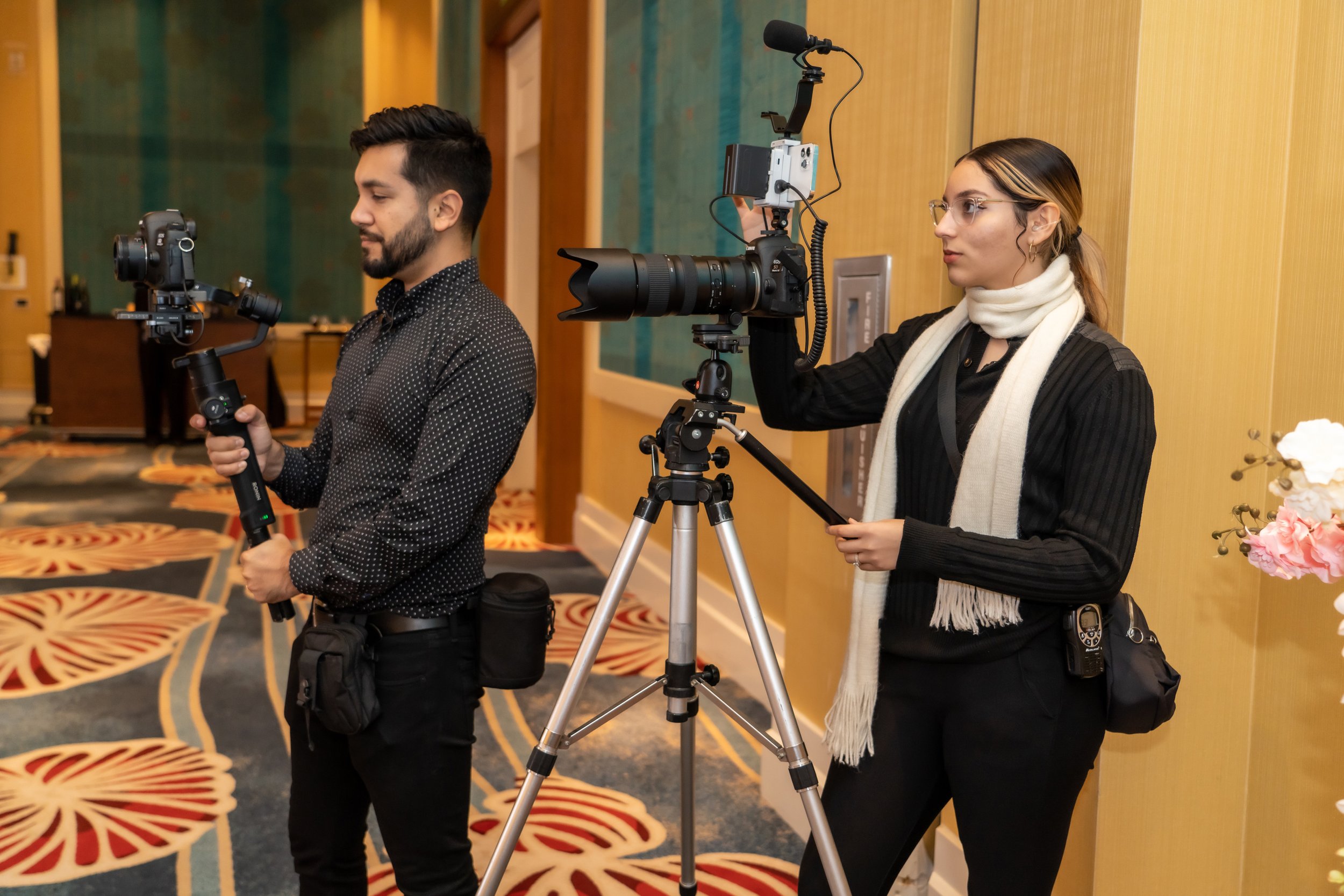 Two photographers are setting up equipment in an interior space with wooden panel walls and a patterned carpet. The man on the left is holding a camera mounted on a stabilizer, and the woman on the right is adjusting a camera on a tripod, wearing glasses, a black jacket, and a white scarf.