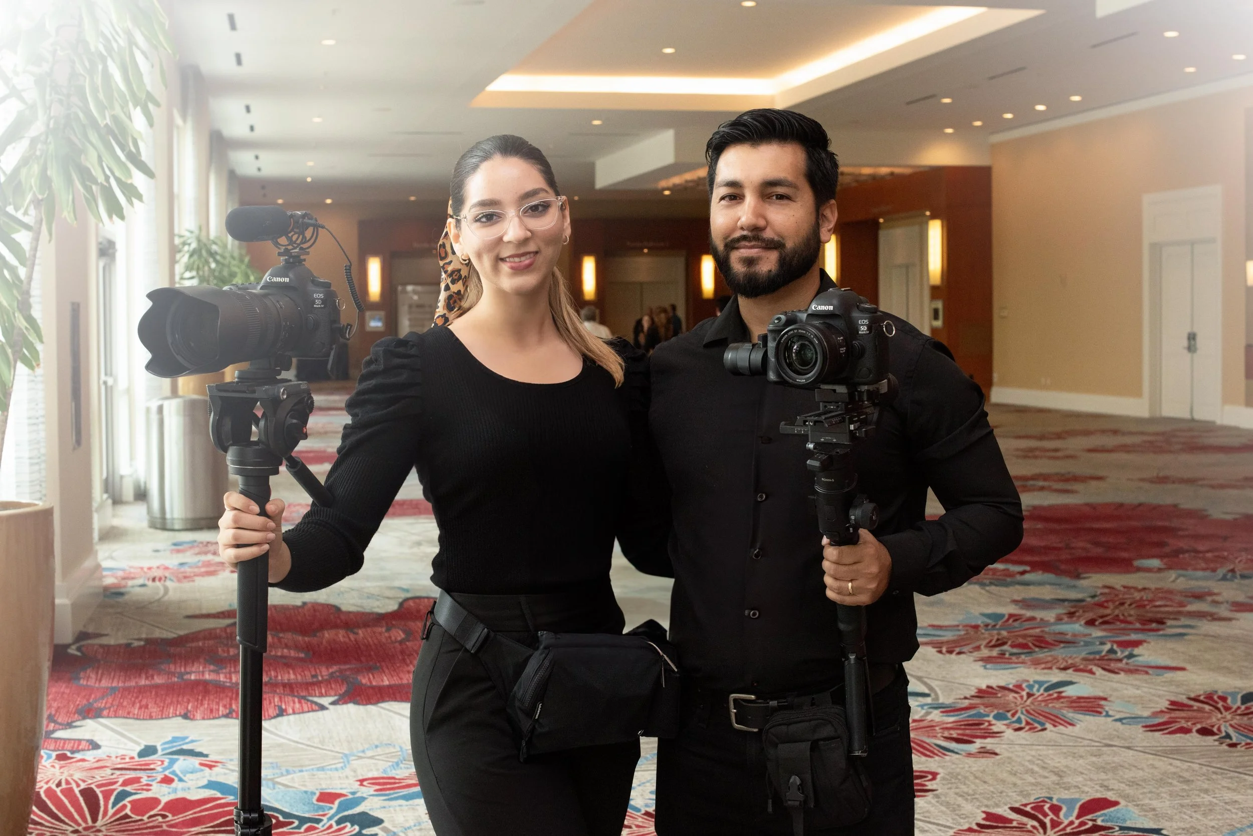 A man and woman smiling with cameras in an indoor setting, possibly a hotel lobby or conference center, with a patterned carpet and plants in the background.