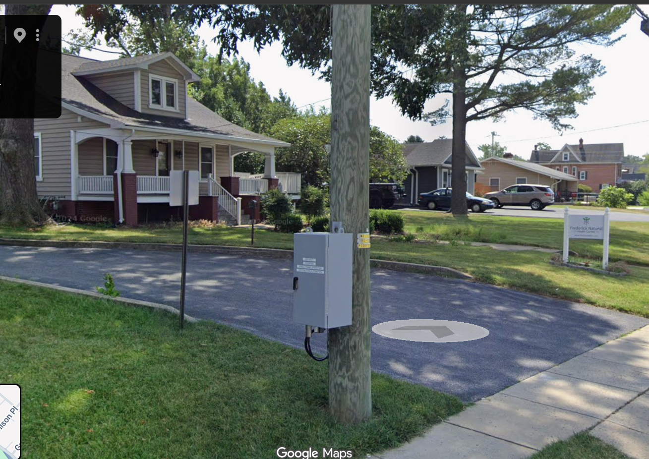 Residential house with a front porch and well-maintained lawn, a large tree, and a street sign. A white sign with green and black text reads 'Frederick Natural Health Center.'