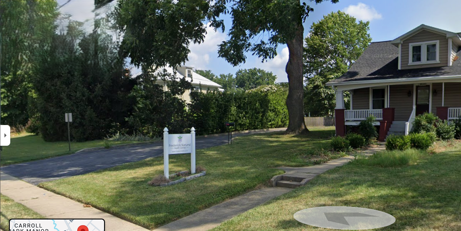 Residential house with a front porch and well-maintained lawn, a large tree, and a street sign. A white sign with green and black text reads 'Frederick Natural Health Center.'