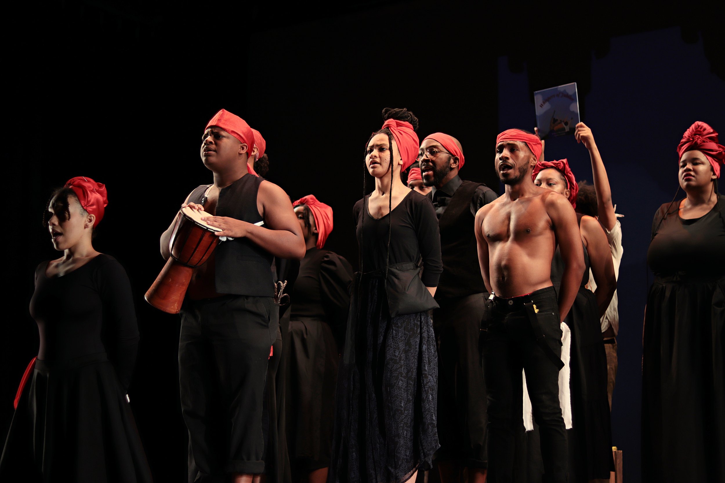 Aurway Artists are standing and singing on stage wearing all black with red head wraps. Marcus Beckett is playing a djembe the most downstage. Taken at a performance of Whispers of Jubilee at Arts High School in Newark, NJ