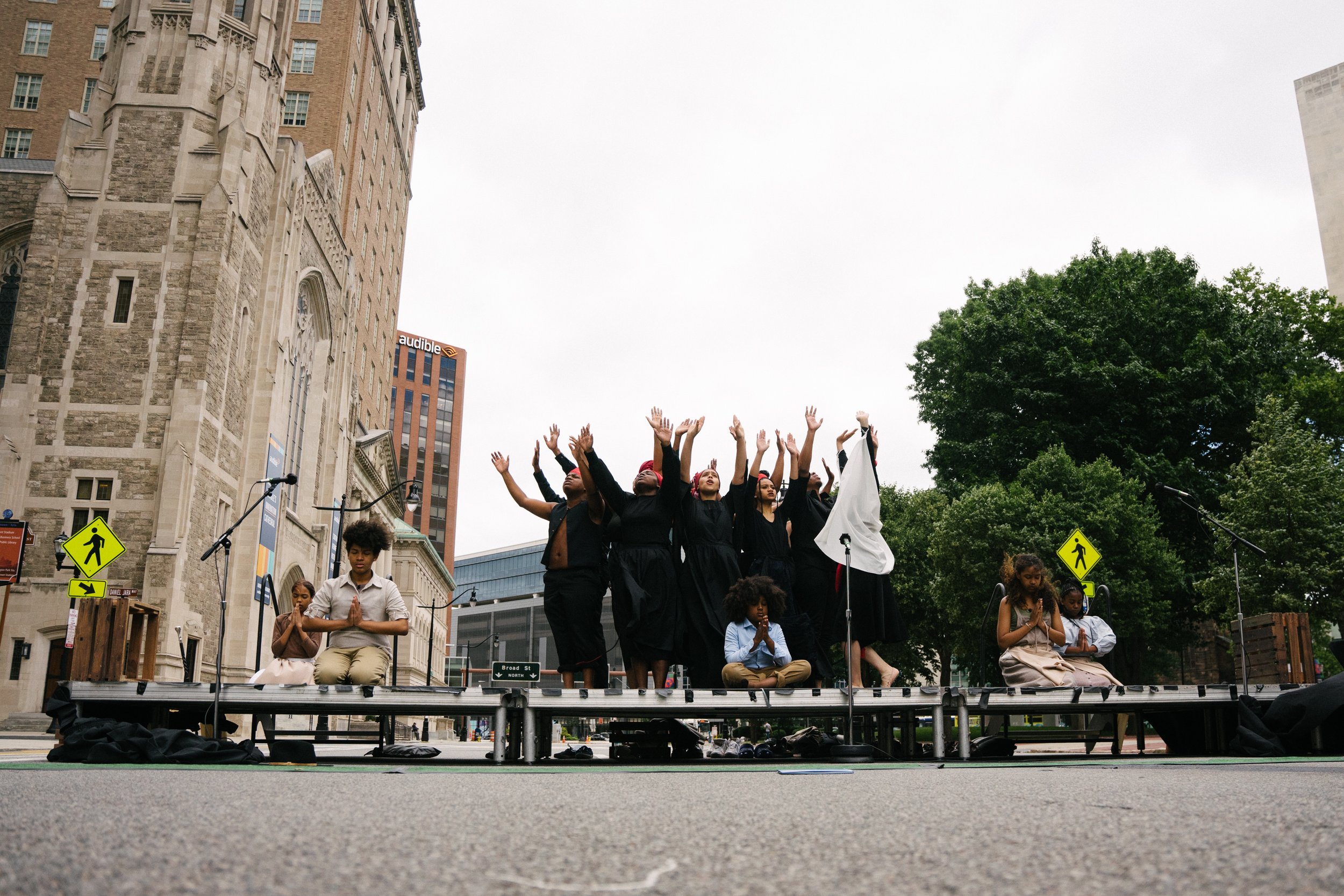 Photo by Bryon Summers
Aurway Artists stand center on a stage outdoors on Washington Street in Newark NJ. Wearing all black, with their arms raised above their heads. At the Newark Museum of Art's Juneteenth Celebration.