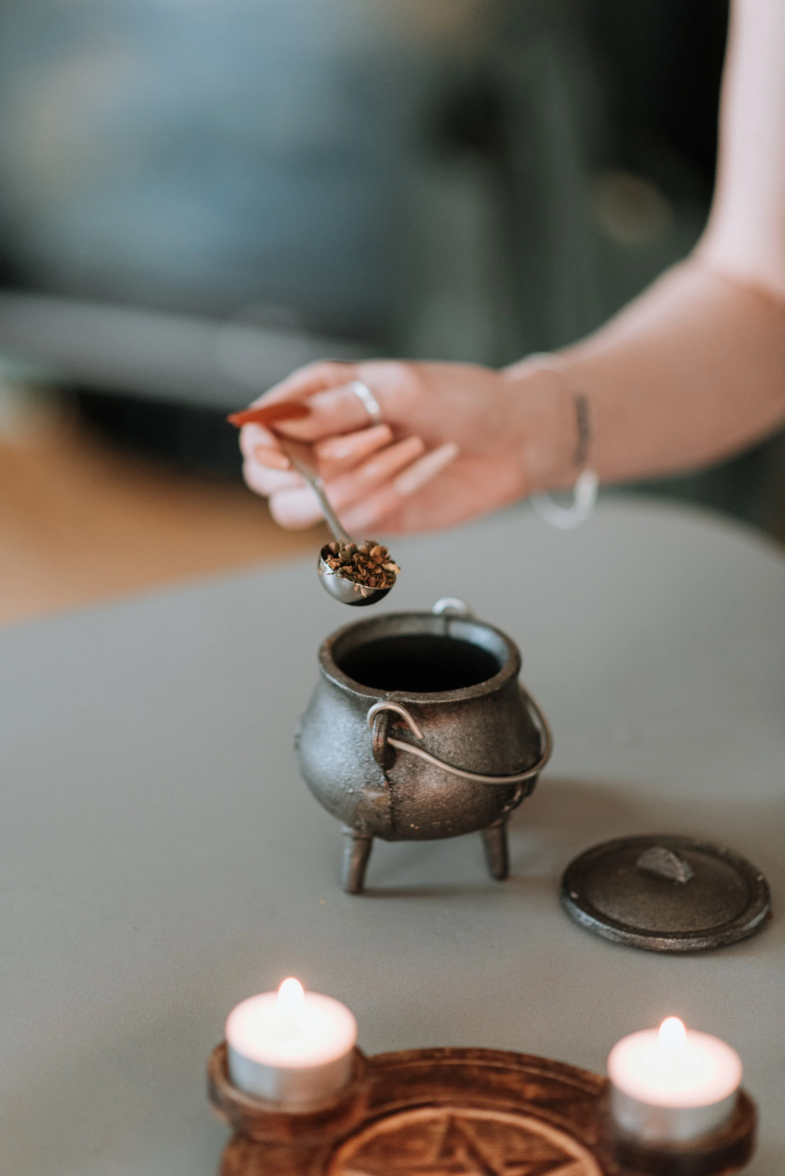 A witch working magic, we see a hand pouring herbs into a cauldron with two candles beside it.