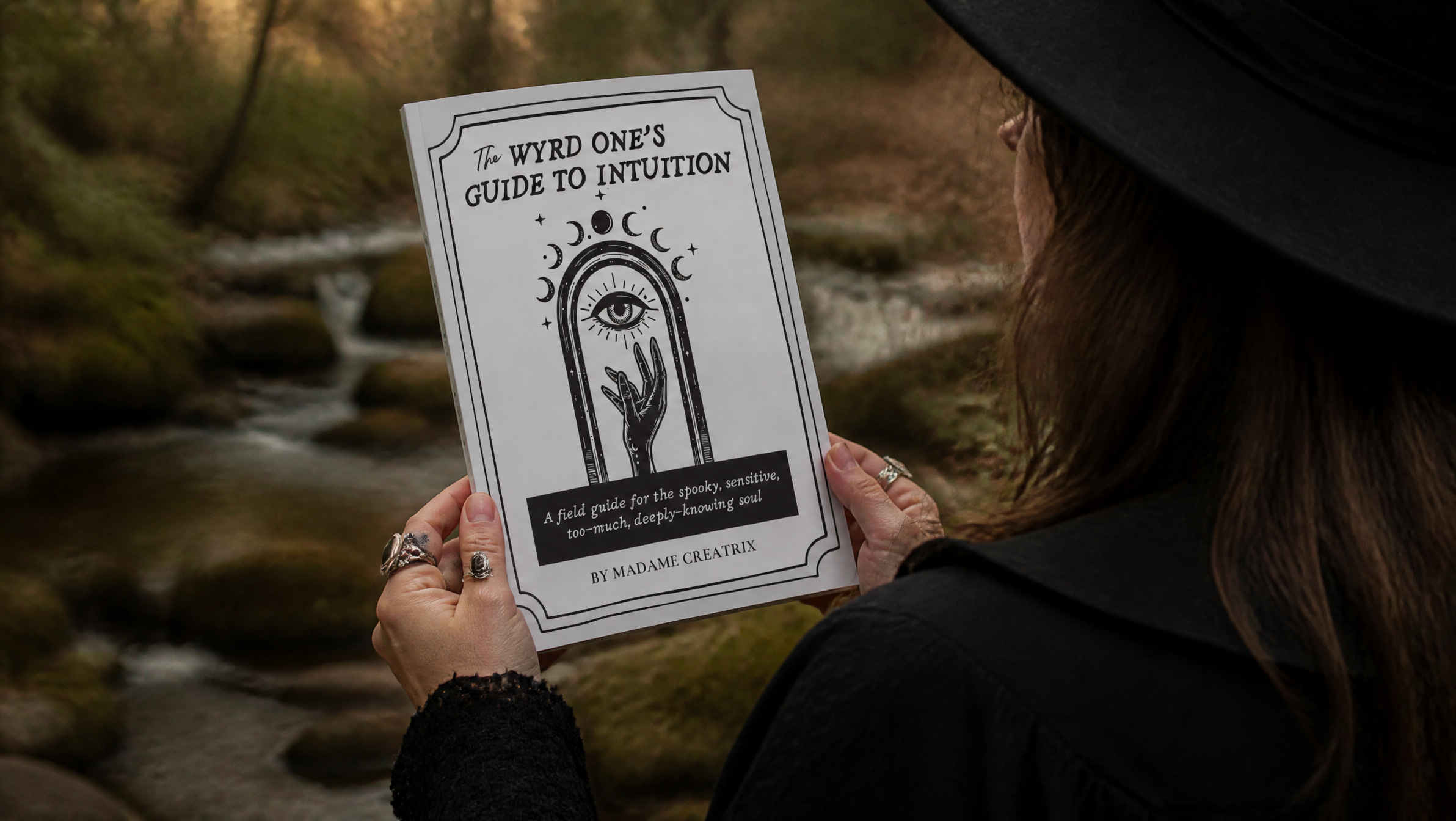 Person in a black witch hat holding a printed copy of The Wyrd One’s Guide to Intuition by Madame Creatrix outdoors near a mossy creek.