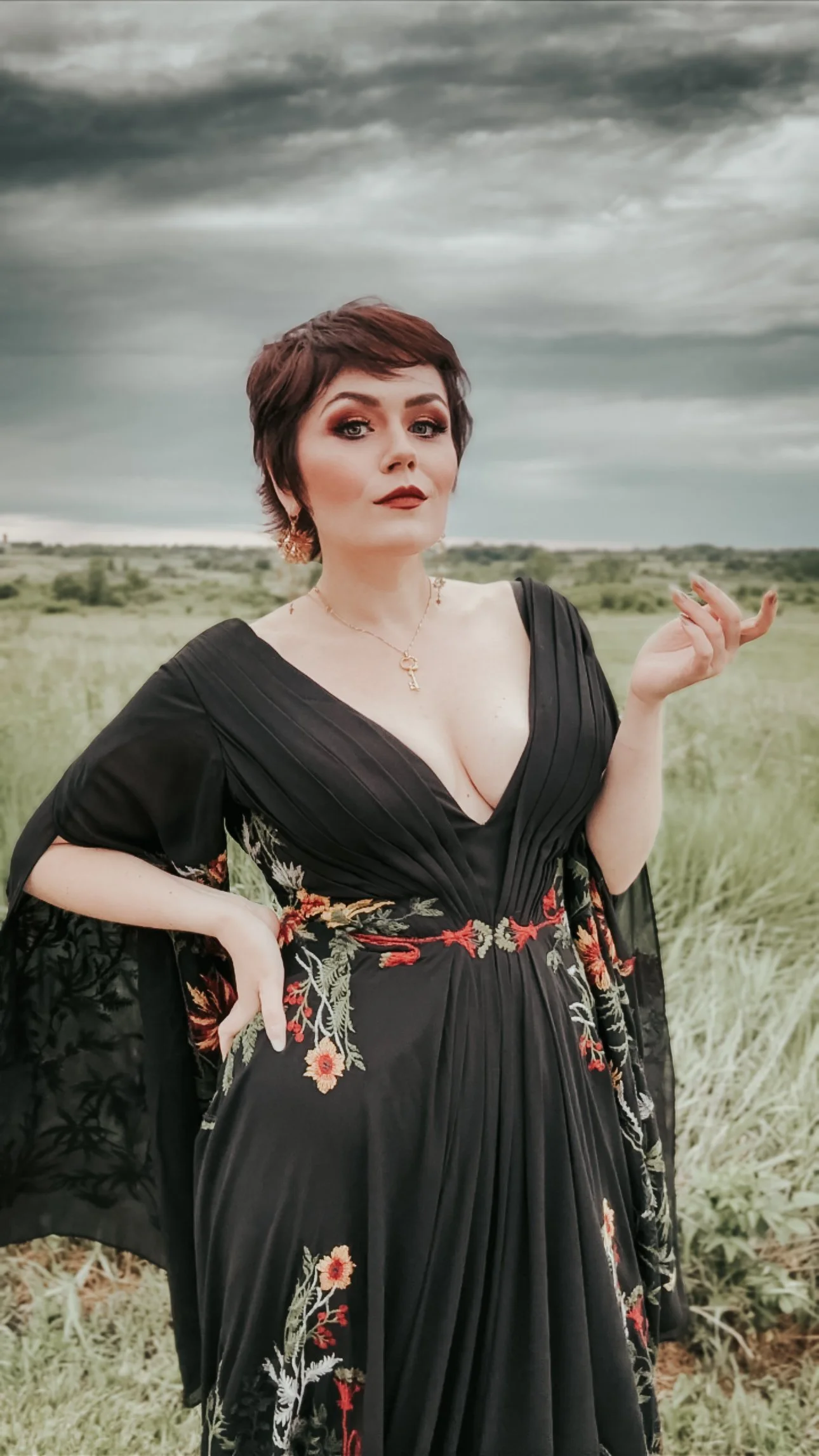A woman with short, dark hair wearing a black dress with floral embroidery, standing outdoors in a field with dark, cloudy skies.