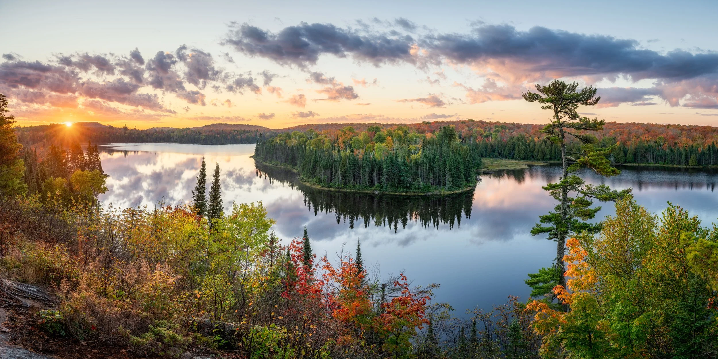 Panoramic view of a river with a small island surrounded by dense forest during sunset, with colorful autumn foliage and partly cloudy sky.