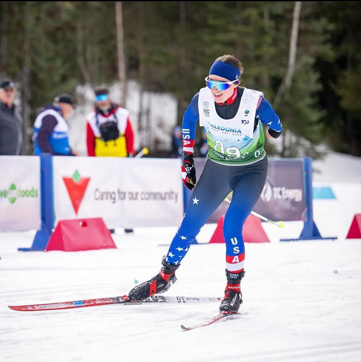 Dani in cross-country skier in USA team gear smiling and skiing on snow during a race, with others and trees in the background.