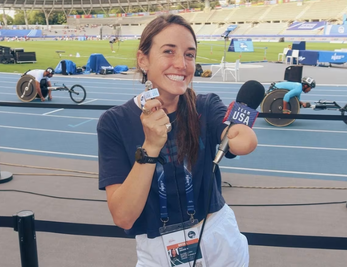 Dani smiling at the camera during a track and field event, holding a bid in one hand and a microphone in the other, with a wheelchair race in progress behind her at a stadium.