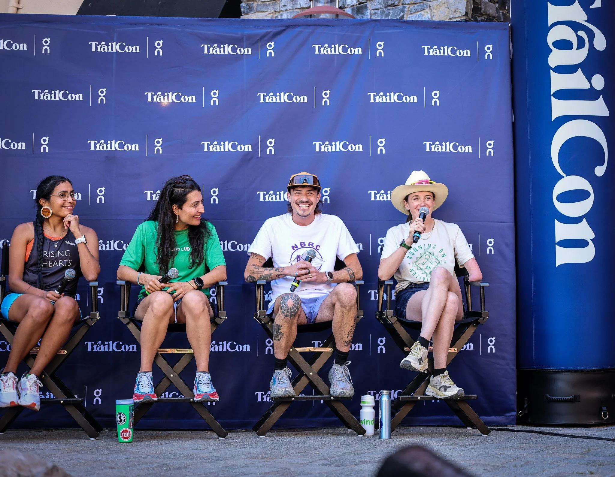 Four people sitting on chairs on a stage with a blue TrailCon backdrop, talking into microphones, with water bottles in front of them, appearing to participate in a panel at an outdoor event.