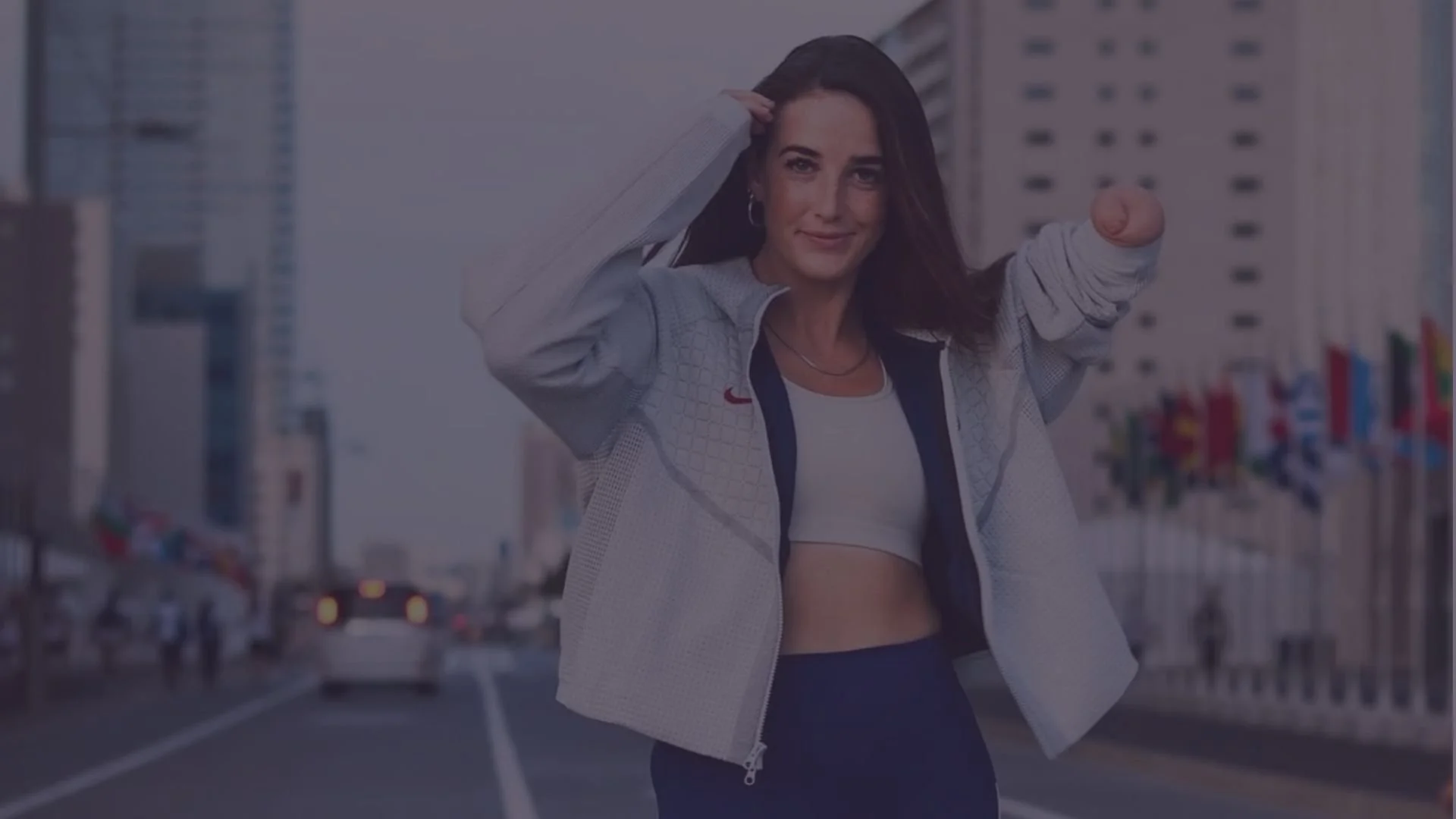 A woman with long dark hair standing on city street, smiling, wearing a sports jacket, white crop top, and black pants, with city buildings and flags in the background.