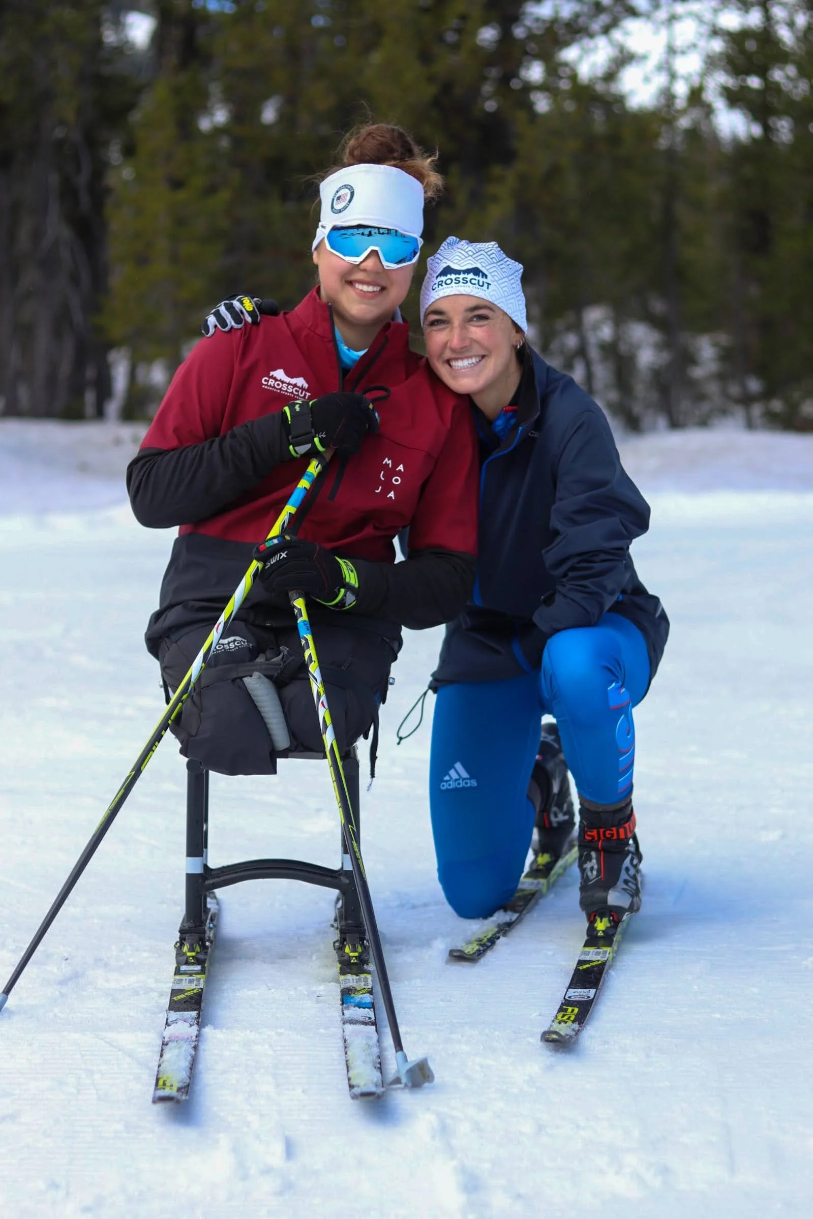 Dani and friend dressed in winter sports gear, smiling, on a snow-covered landscape with trees in the background. One woman is on adaptive skis, and the other is standing beside her.