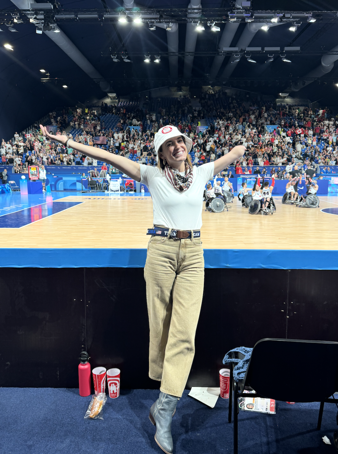 Dani smiling with arms outstretched at a wheelchair basketball event on an indoor basketball court, with a large crowd in the stands.