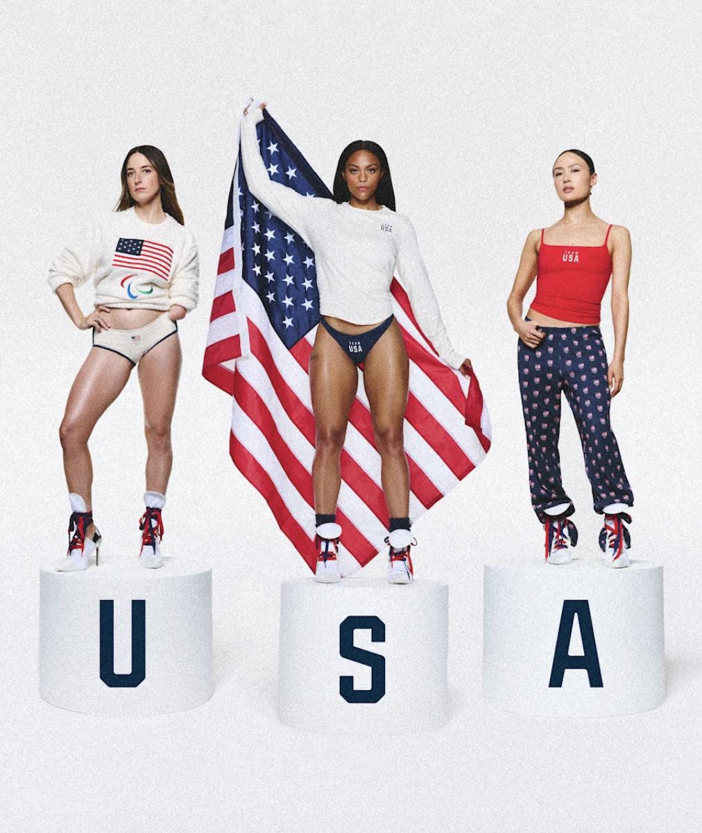 Three women standing on podiums with letters spelling U-S-A, with the woman in the center holding an American flag.