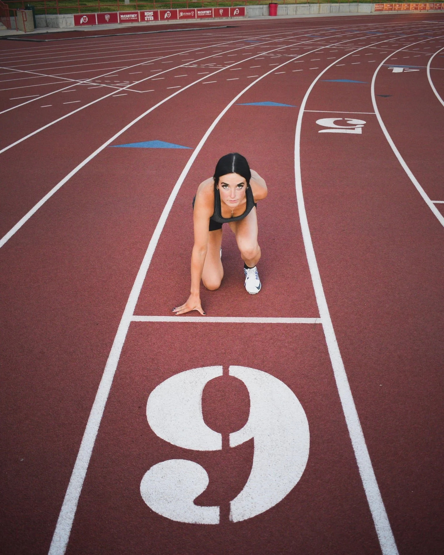 Dani dressed in athletic clothing in a starting position on a red running track marked with white lanes and numbers, preparing for a race.