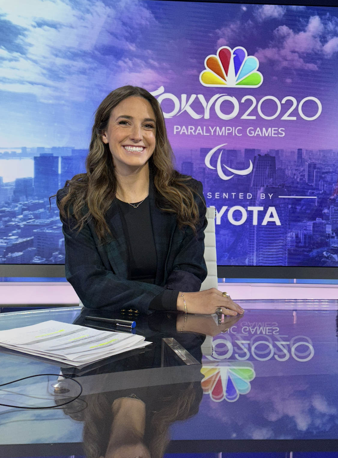 Dani with long brown hair, smiling, sitting at a news desk with papers and a pen, in front of a large screen displaying the Tokyo 2020 Paralympic Games logo and branding.