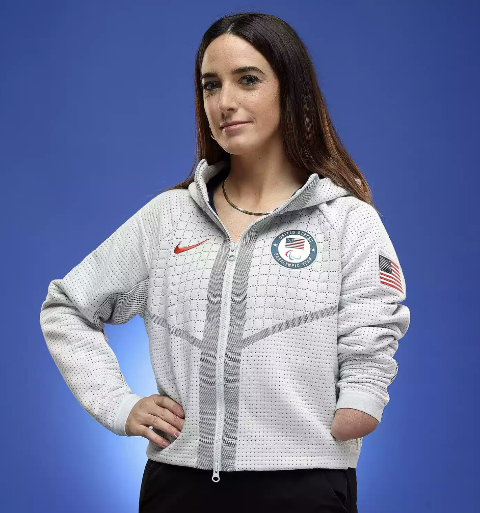 Dani with long brown hair wearing a gray USA team jacket with Olympic and American flag patches, standing against a blue background.