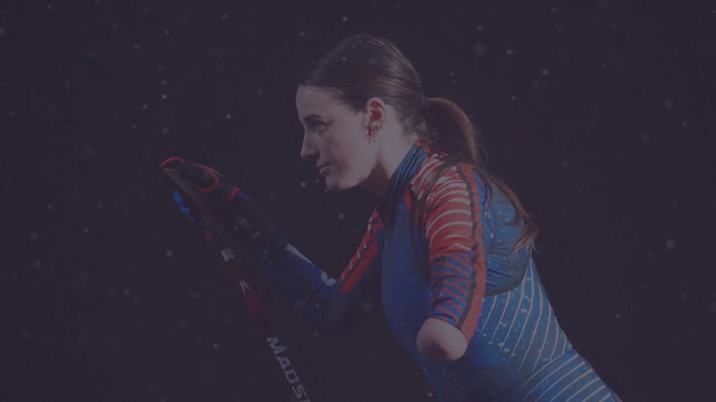 A woman professionally dressed in sportswear holding a ski pole, with snow falling around her in the dark.