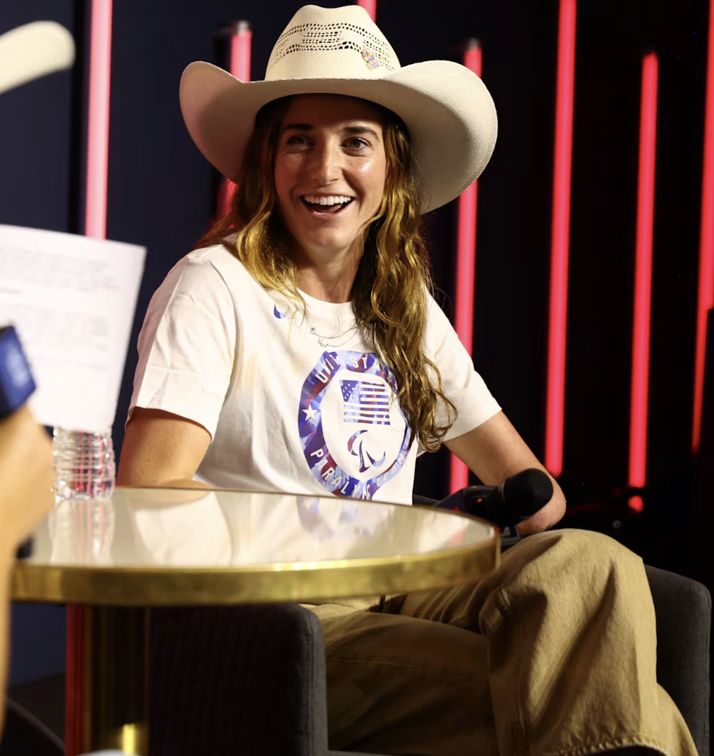Dani in a large cowboy hat and casual clothing is smiling during an event, sitting on a chair with a microphone in her hand, with a dark background and neon pink vertical lights.