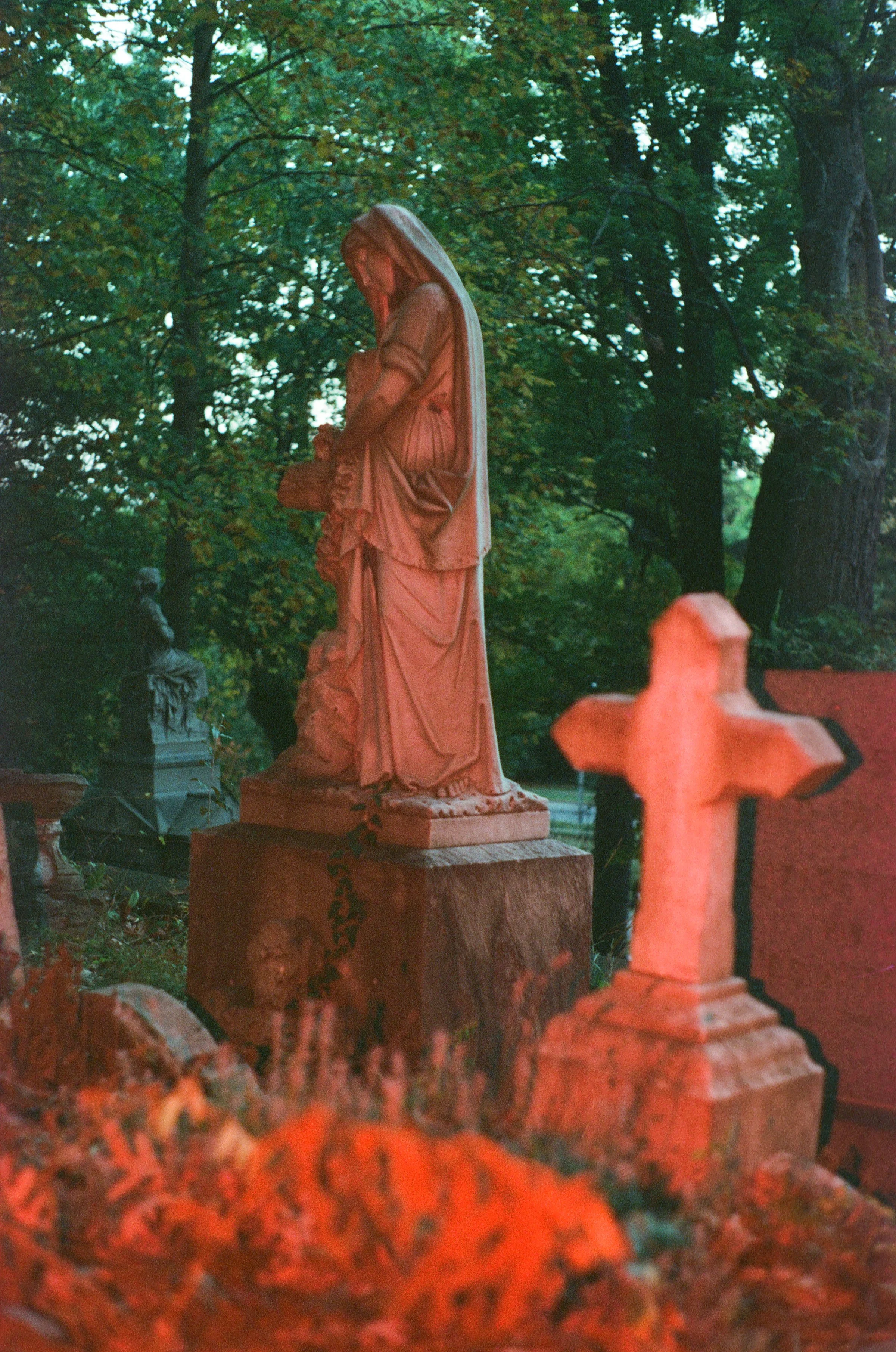 A sculpture of Jesus Christ with long hair and a beard, standing on a stone pedestal in a park with trees in the background.
