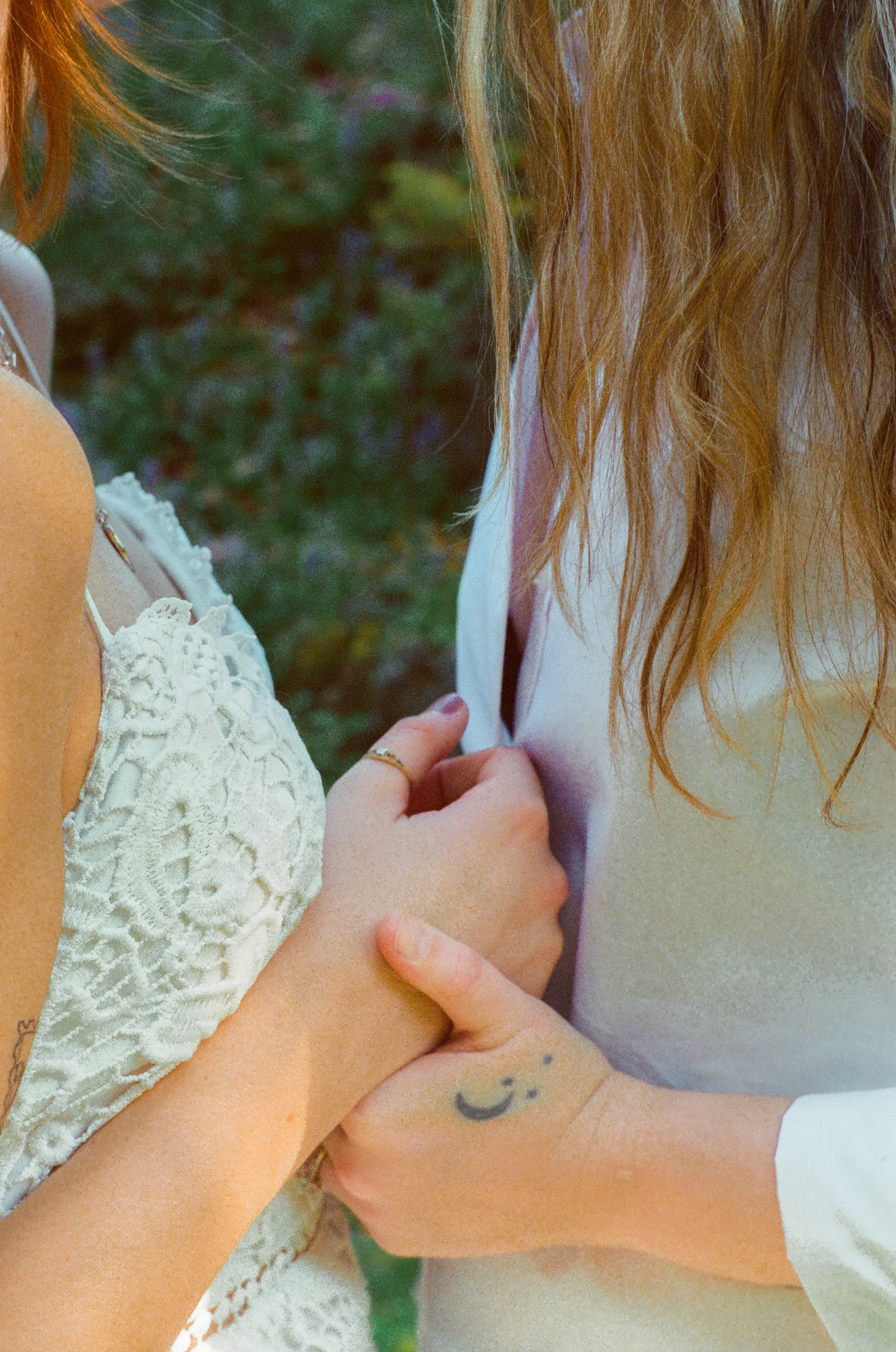 Two women are close together, holding each other's arms, one wearing a white lace top and the other a light-colored sleeveless garment. The woman on the left has orange hair and visible jewelry, while the woman on the right has long, wavy red hair. T