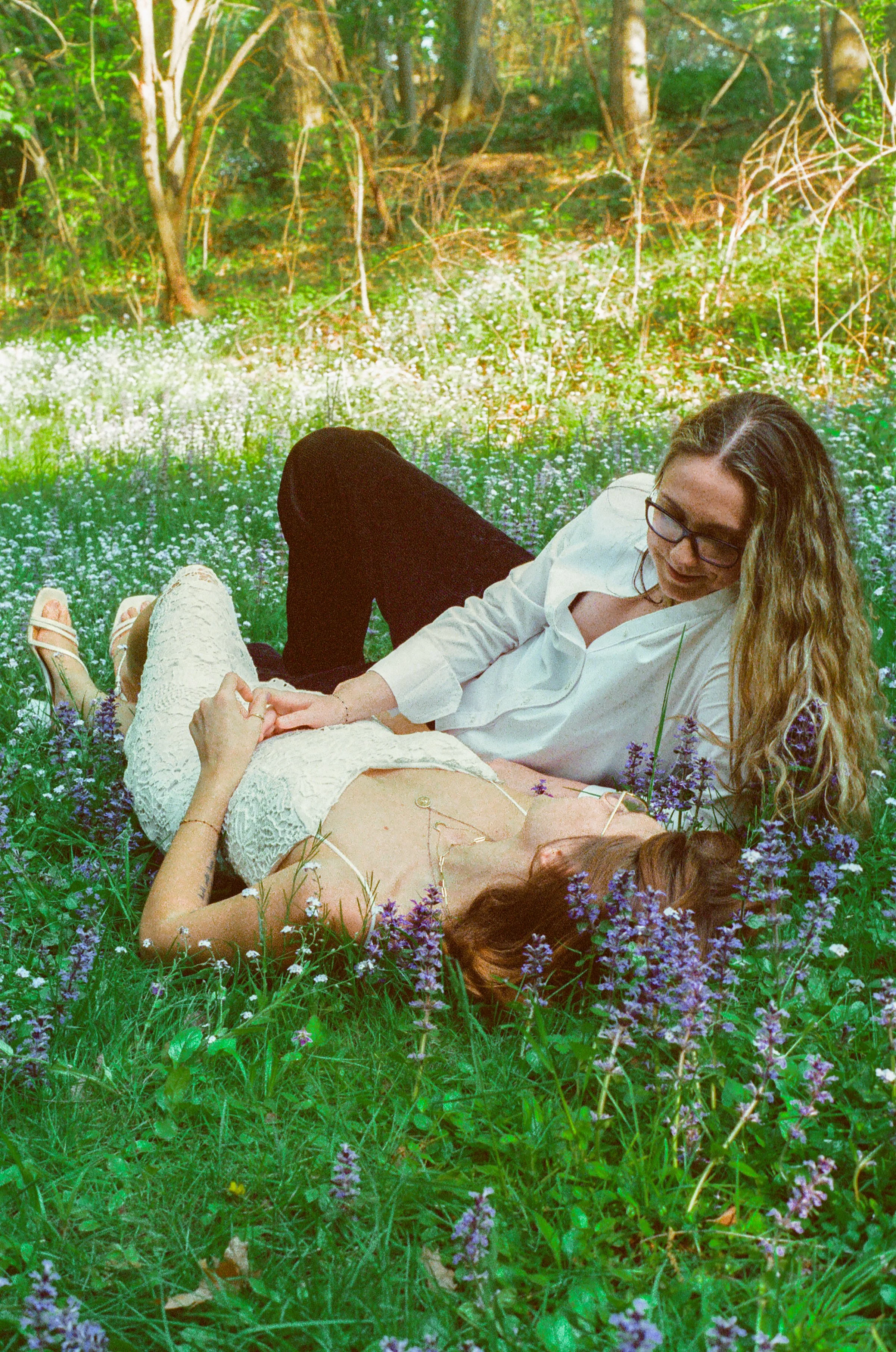 A woman and a man lying in a field of purple flowers with a background of trees and greenery.