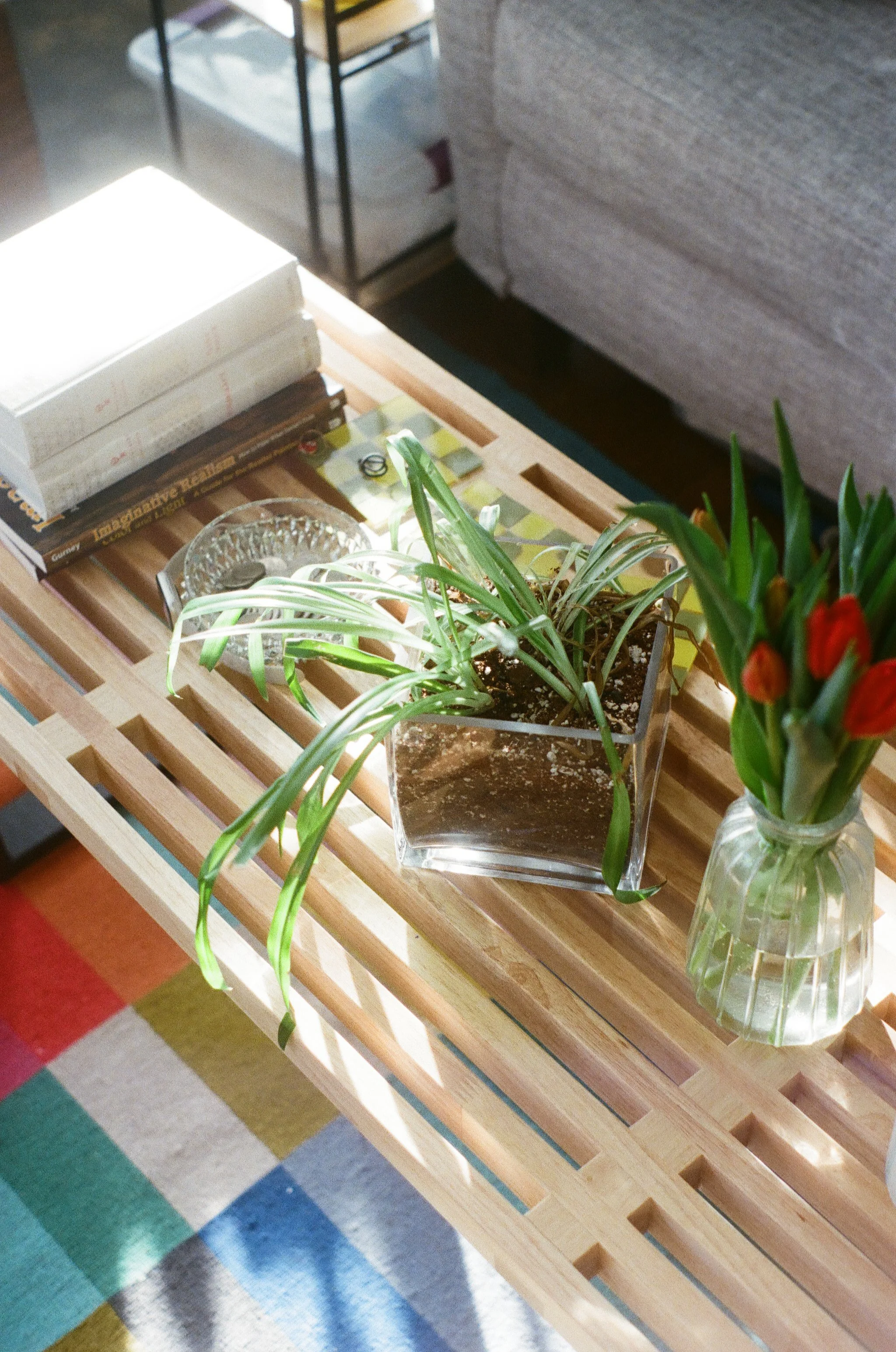 A wooden slatted table with a clear glass vase holding green leafy plants and red flowers, a small dish, and some books, with a colorful tiled floor underneath.