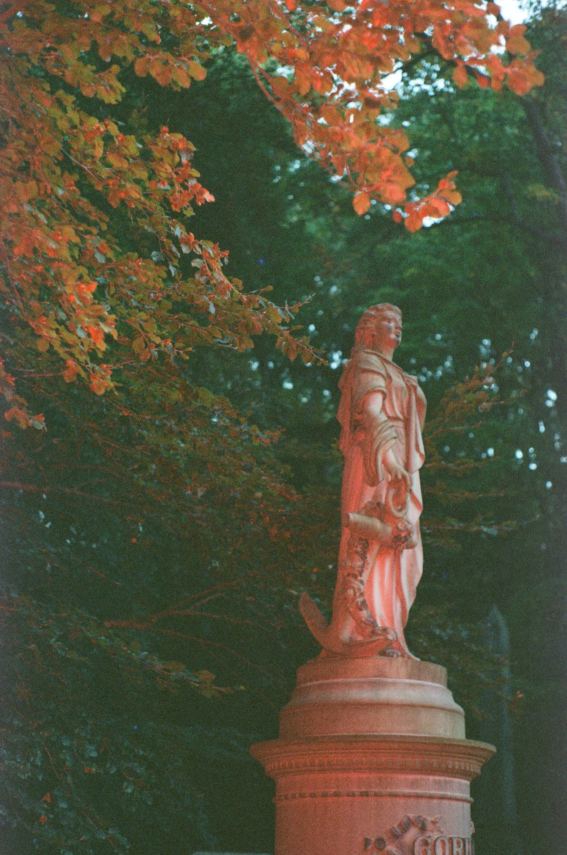 Statue of a woman with flowing hair and detailed clothing, standing on a circular base, surrounded by trees with red and green leaves.